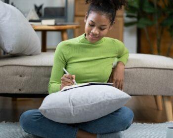 Young woman journaling in a cozy living room, sitting cross-legged on the floor with a pillow and notebook, practicing mindfulness and gratitude journaling.