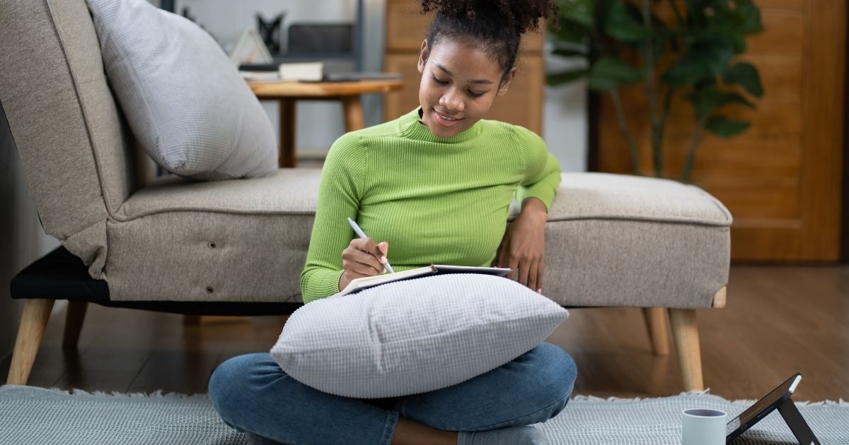 Young woman journaling in a cozy living room, sitting cross-legged on the floor with a pillow and notebook, practicing mindfulness and gratitude journaling.